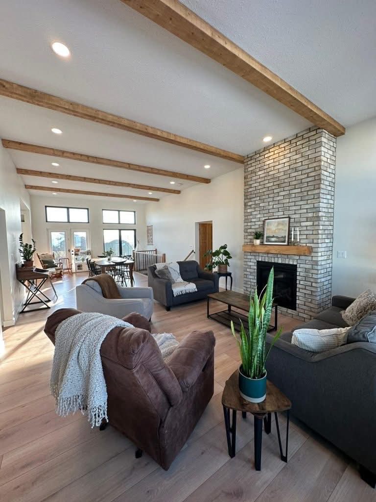 Spacious living room with wooden beams, a brick fireplace, and cozy seating. Soft lighting and indoor plants create a warm, inviting atmosphere.