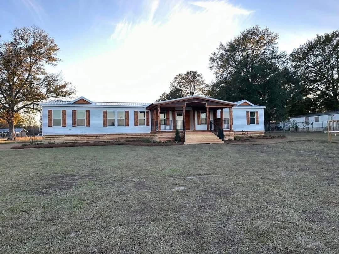 A white, single-story modular home with a wooden porch stands in an open, grassy yard. Tall trees border the property under a clear blue sky.