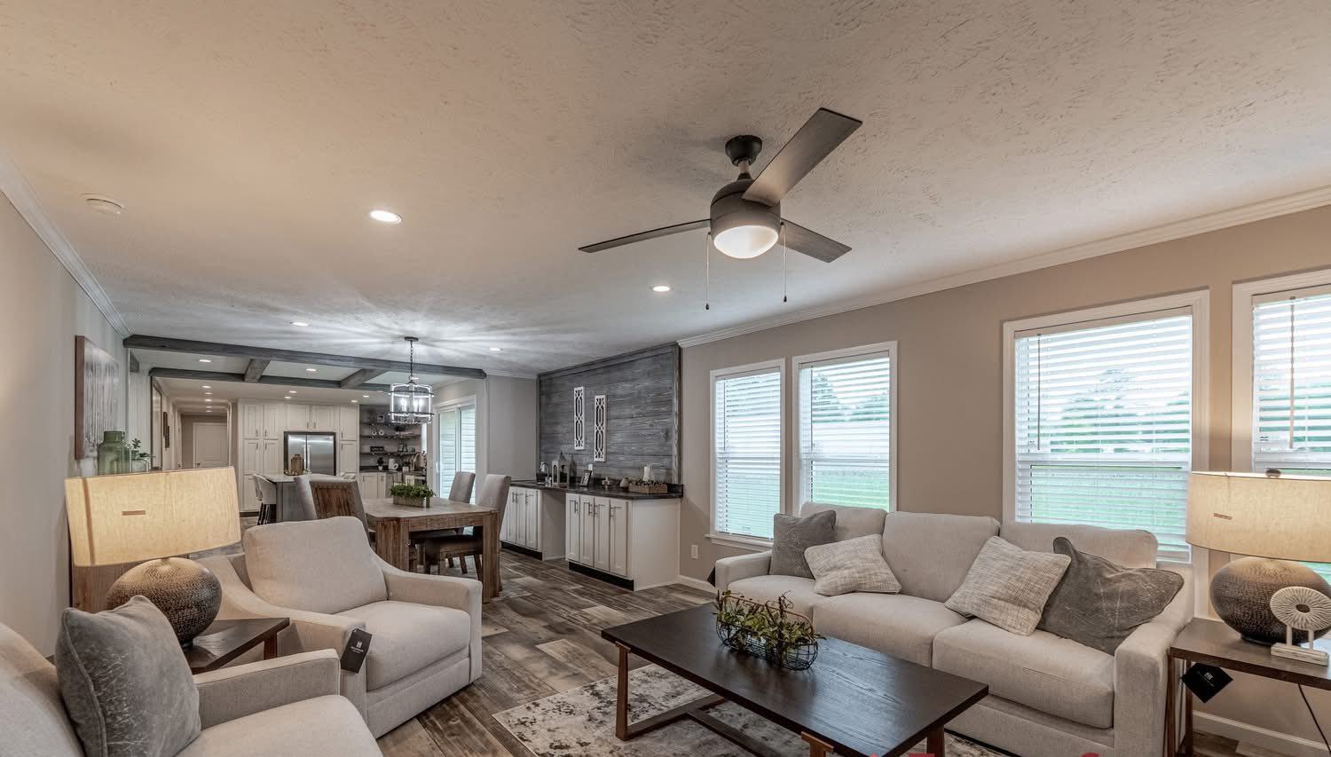 Modern living room with neutral tones features a ceiling fan, plush sofas, and a wooden coffee table. Large windows offer natural light, creating a cozy, inviting atmosphere. Dining area and kitchen are visible in the background.