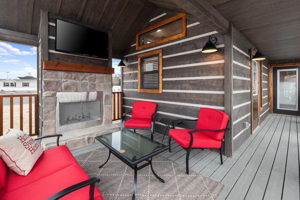Cozy outdoor patio with dark wood walls, stone fireplace, red cushioned chairs, glass table, and decorative pillows. Warm, inviting, and modern.