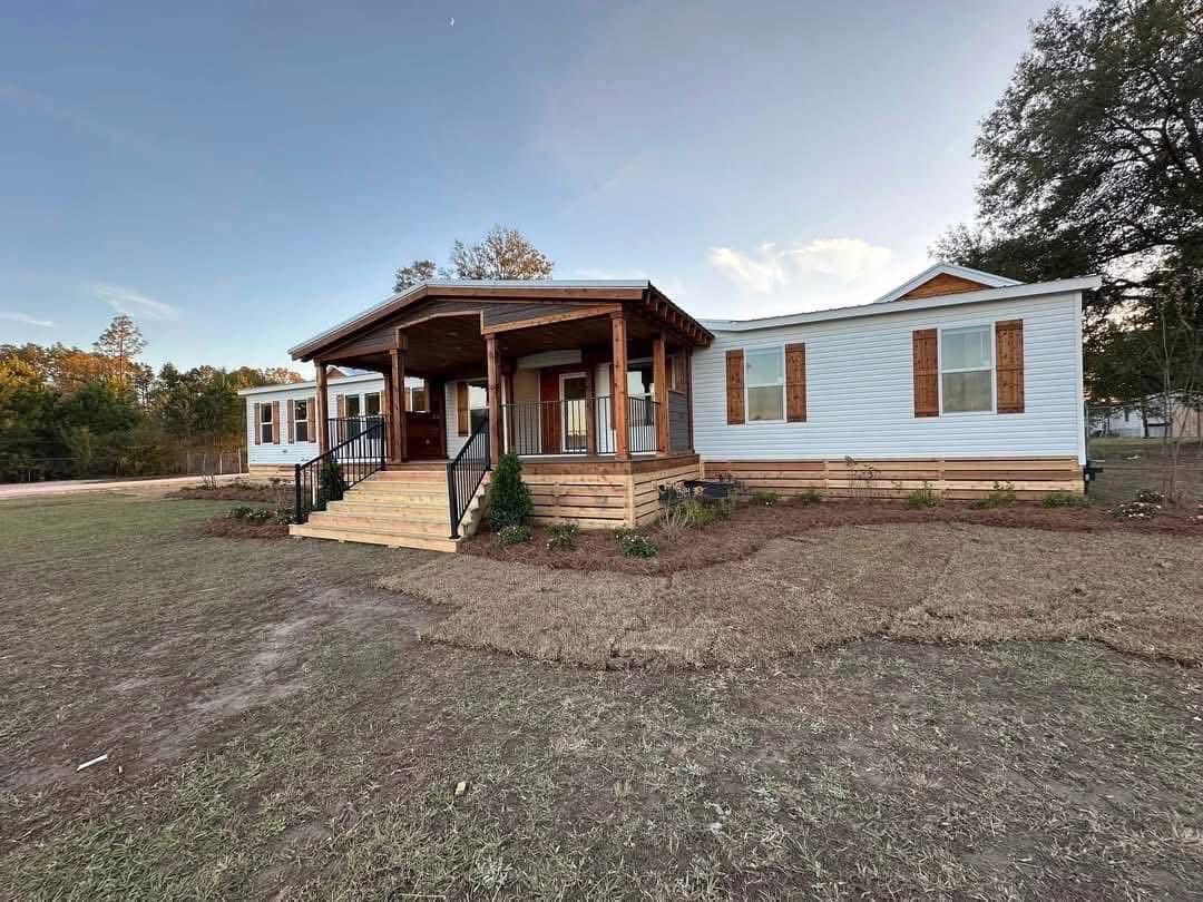 A white modular home with a wooden porch and steps sits on a grassy lawn. Trees and a clear sky are in the background, creating a serene rural setting.
