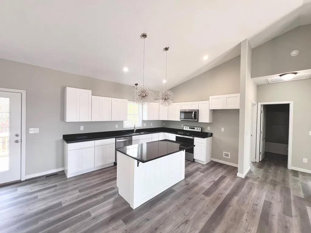 Modern kitchen with sleek white cabinets, black countertops, and center island. Stainless steel appliances, pendant lights, and gray flooring create a clean, airy feel.