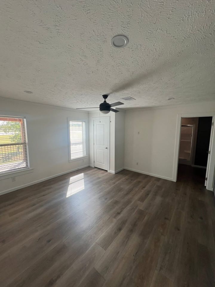 Empty room with light wood flooring and white walls, featuring a ceiling fan. Sunlight streams through two windows, creating a bright, airy feel.