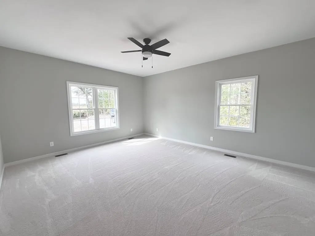 Spacious, empty room with light gray walls, white carpet, two large windows, and a ceiling fan. Abundant natural light creates a calm atmosphere.