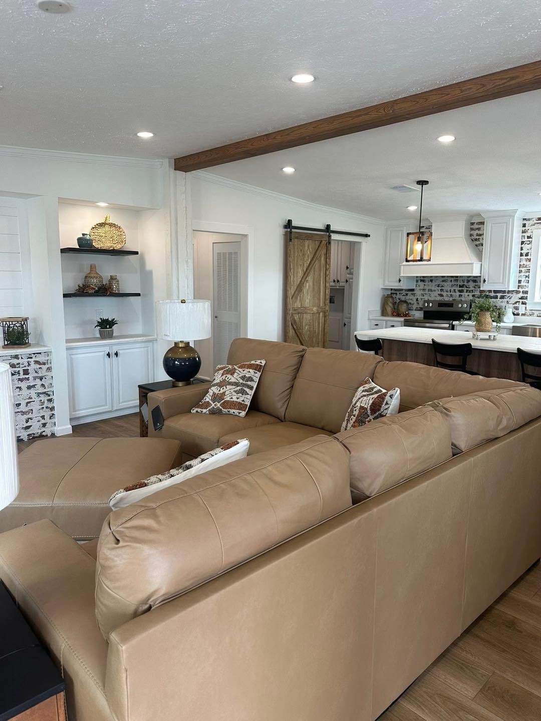 Modern living room with a large tan sectional sofa, accent pillows, and cozy lighting. Open kitchen features white cabinets and a rustic barn door.