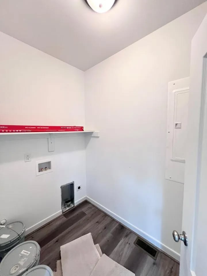Narrow laundry room with white walls, wooden floor, and overhead light. Shelf with paint cans, wall hook-ups, and a gray vent on the left. Calm ambiance.