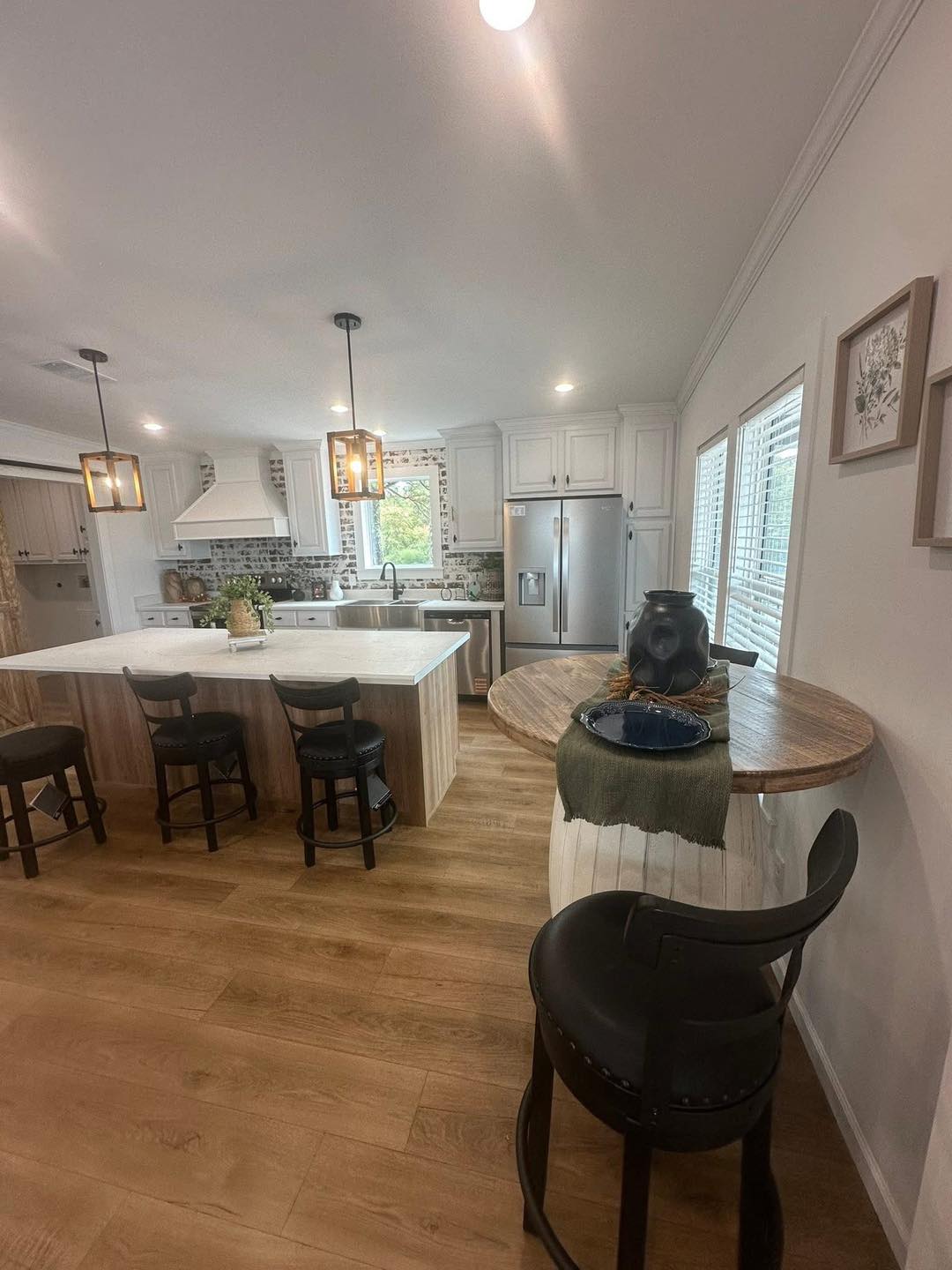 A modern kitchen with light wood flooring, a central island with four black chairs, pendant lighting, white cabinetry, stainless steel appliances, and a small round table with black stools, conveying a warm, inviting atmosphere.