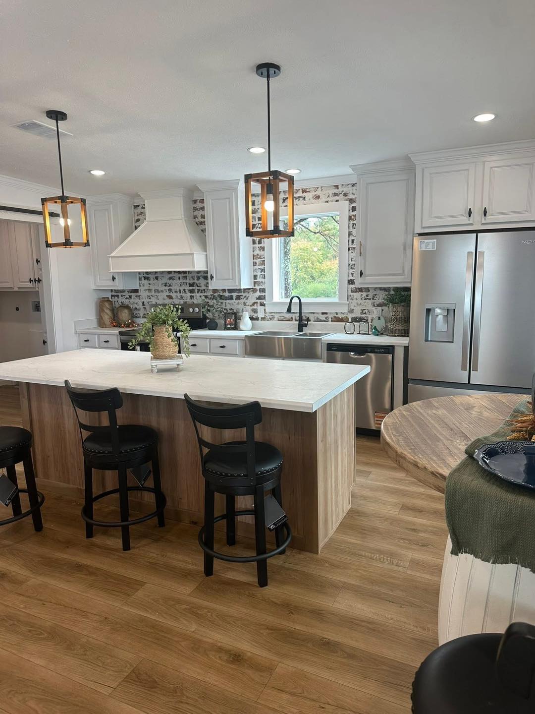 Modern kitchen with wooden floors, white cabinets, and a central island featuring three black stools. Pendant lights hang above a white countertop.