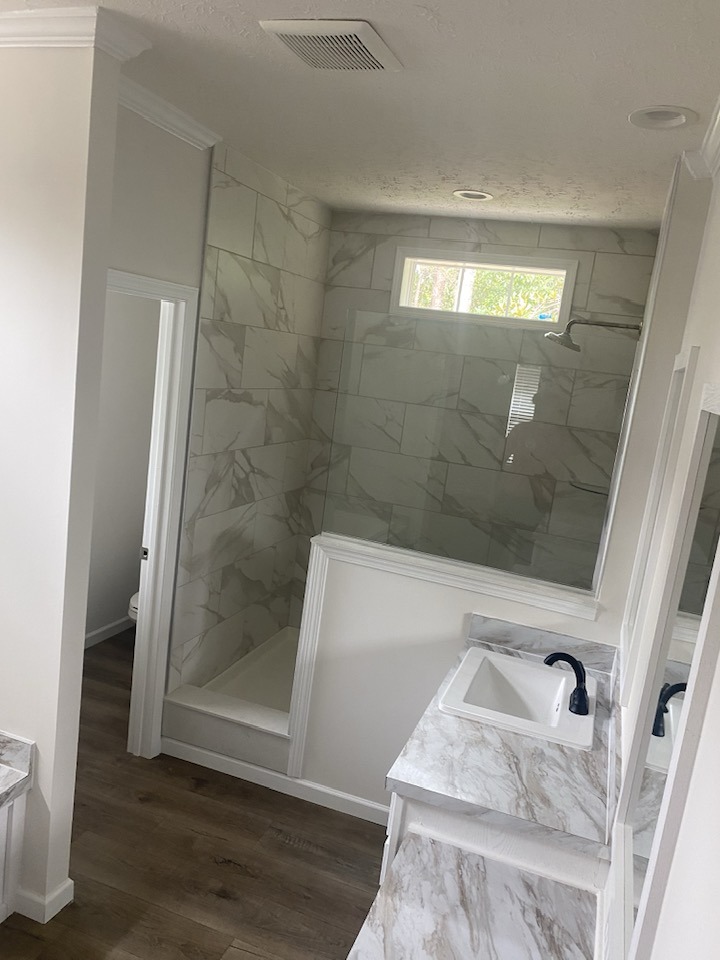 Modern bathroom with marble-patterned shower and countertop, white sink, and black faucet. Light wood floor and natural light create a calming atmosphere.