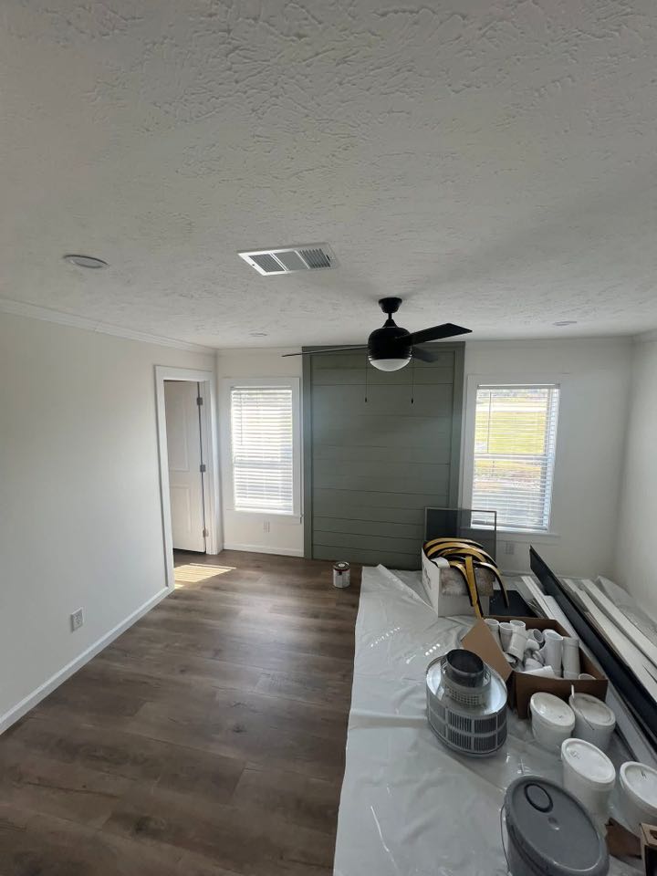 A room under renovation with white walls, wood floor, a ceiling fan, and windows. Boxes and paint cans on a table covered in plastic. Calm ambience.