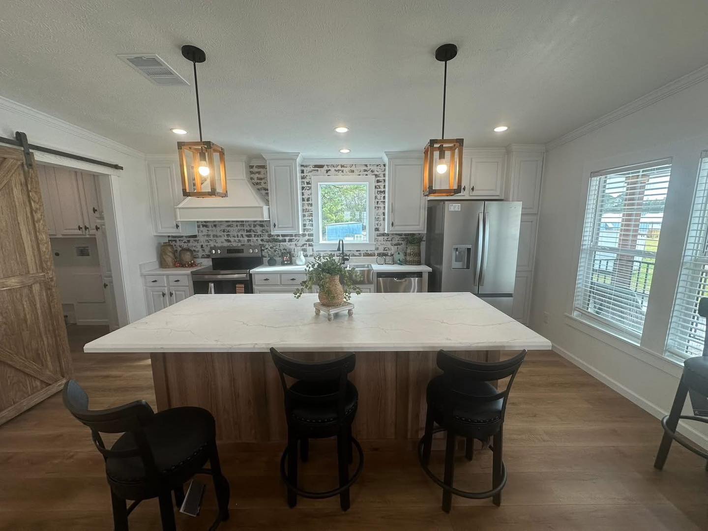 Sleek kitchen with marble island, four black chairs, and rustic pendant lights. White cabinets, brick backsplash, steel appliances, and large window. Cozy and modern.