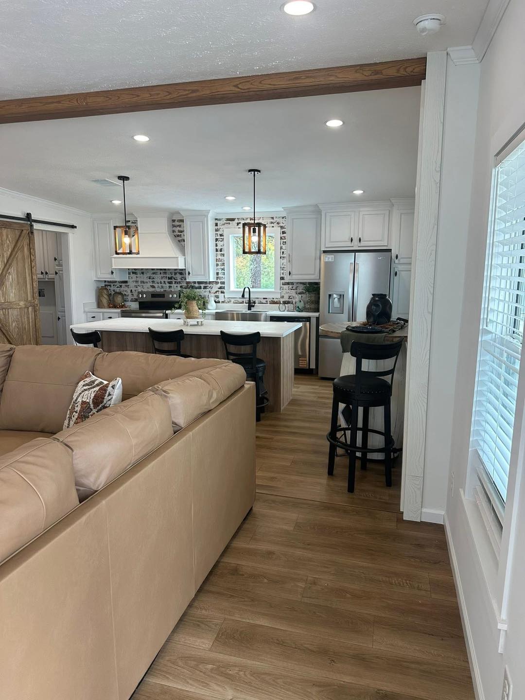Modern kitchen with an island, white cabinets, and hanging lights. A tan sectional sofa and wooden floors add warmth, creating an inviting atmosphere.