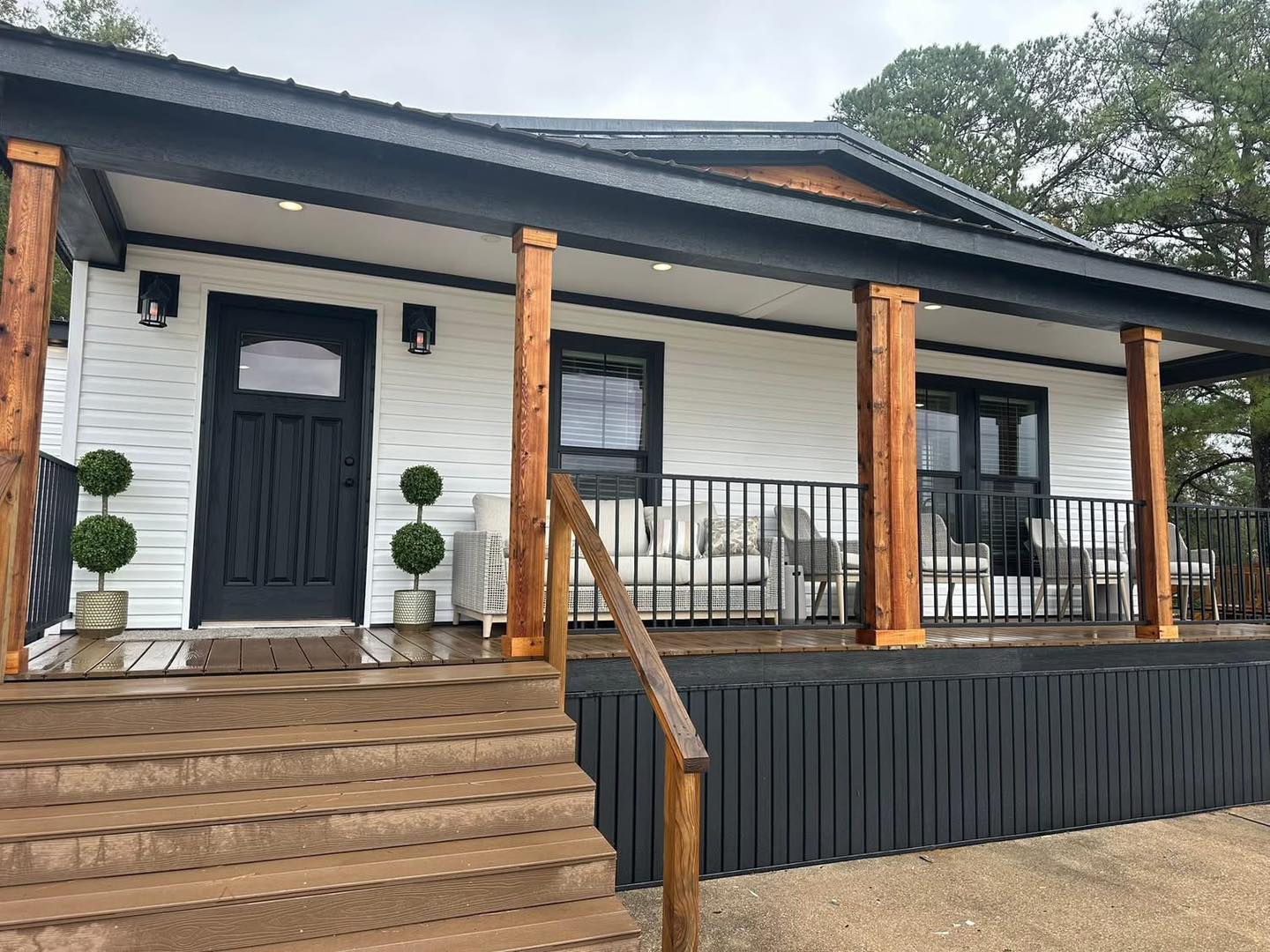 A modern porch with wooden columns and railing, leading to a dark door on a white house. Green potted plants and cozy seating create a welcoming vibe.