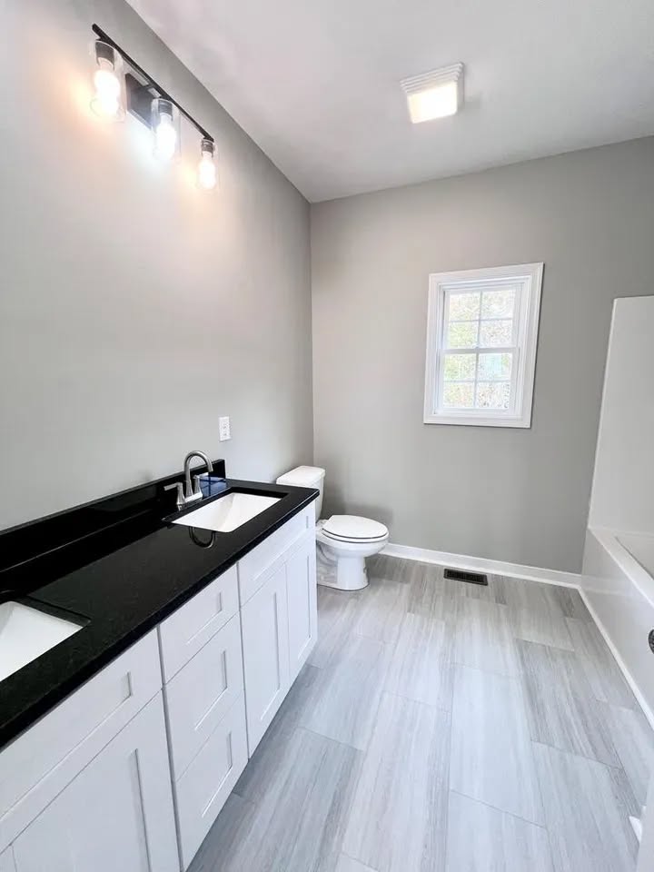 Bright, modern bathroom with light gray walls and floor tiles. Features a black countertop with white cabinets, under a sleek three-light fixture.