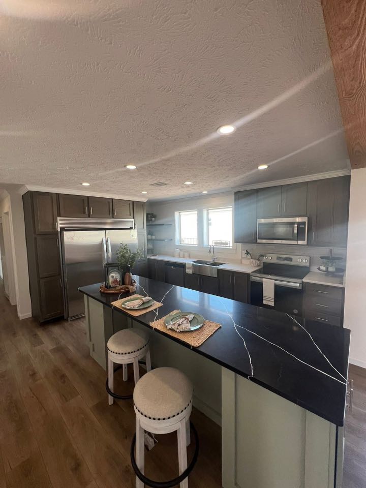 Modern kitchen with a large black island countertop, two cushioned stools, and stainless steel appliances. Bright, airy feel with wood flooring.