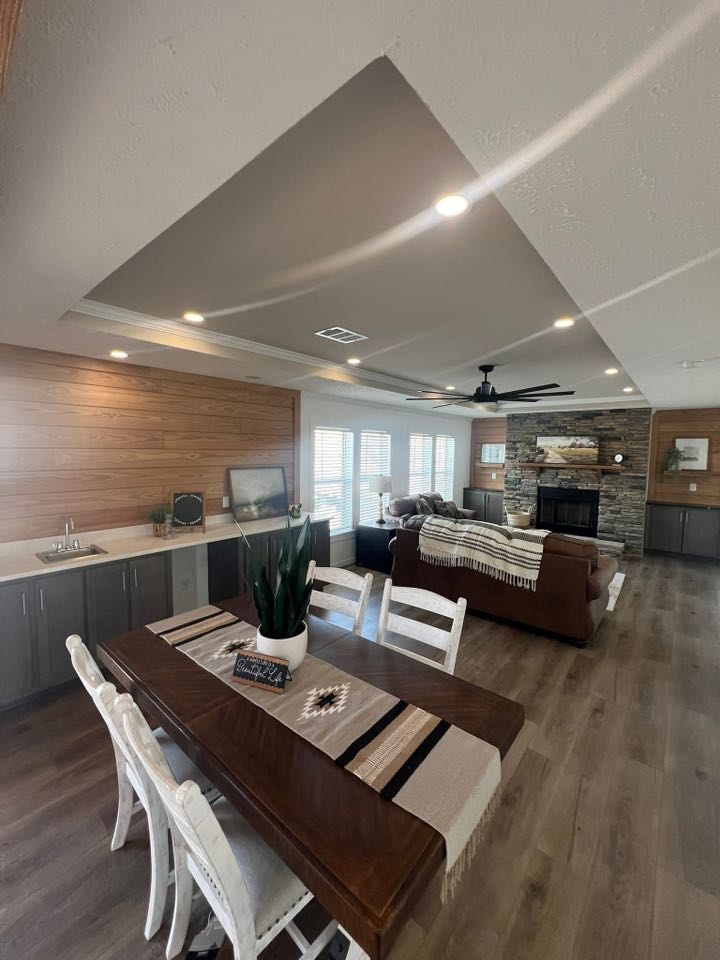 Spacious living room with wooden floors, a dining table with white chairs, a stone fireplace, brown sofas, and large windows offering natural light.