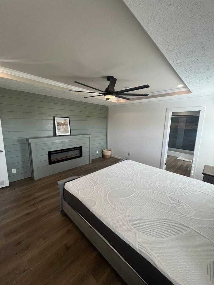 Spacious bedroom with a modern decor, featuring a large bed, wooden floors, a green accent wall, ceiling fan, and a fireplace with a framed picture.