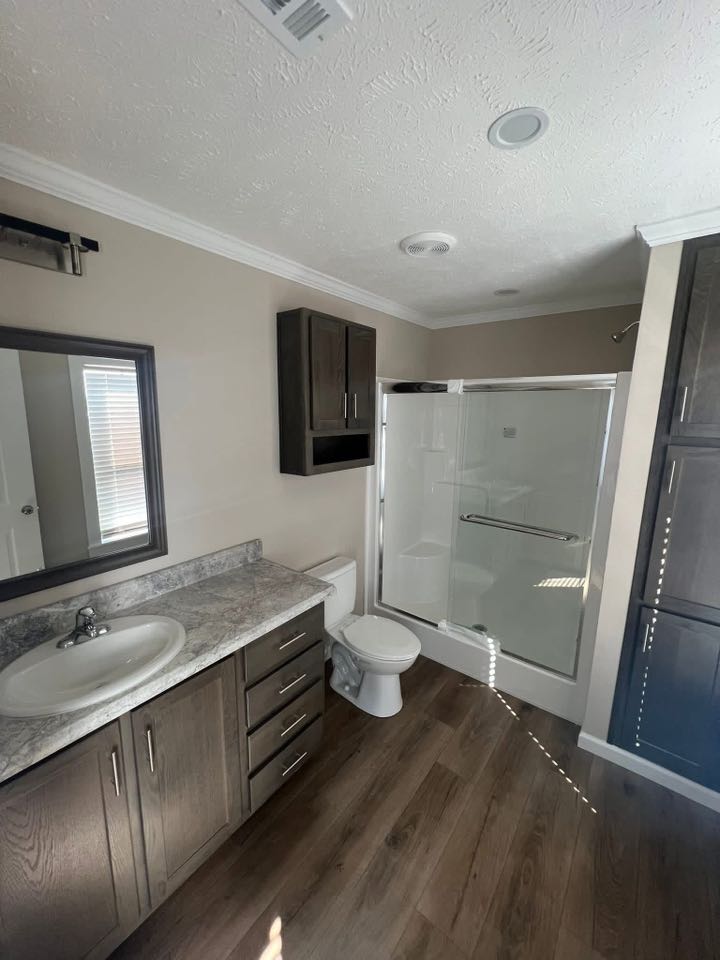 Modern bathroom with wood floors, featuring a marble countertop with a sink, dark wood cabinetry, toilet, and a glass-enclosed shower. Bright natural light.