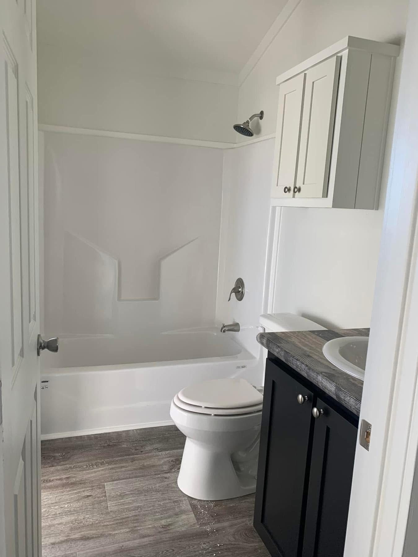 Small modern bathroom with white bathtub, showerhead above, and toilet. Dark vanity with sink, granite countertop. Wood-style flooring.