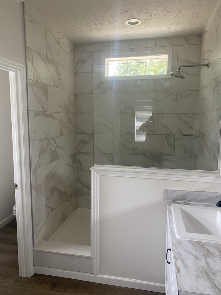 A modern bathroom with a marble-tiled walk-in shower, glass screen, and bright upper window. A white countertop basin is partially visible. Clean, sleek design.