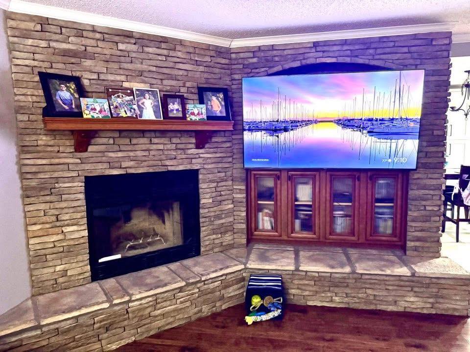 A cozy living room with a stone fireplace and a large TV above wooden cabinets. Photos on a shelf, and a serene sunset image displayed on the screen.