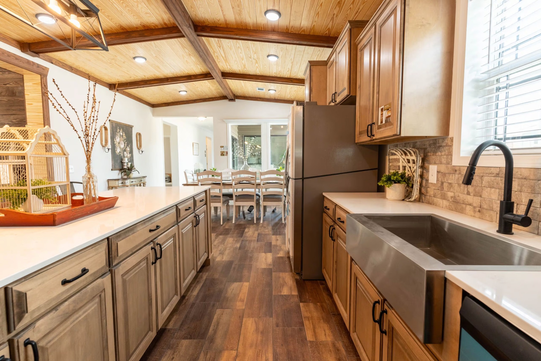 Warm, rustic kitchen with wooden cabinets, stone backsplash, and farmhouse sink. Bright dining area visible, wooden ceiling beams, cozy ambiance.