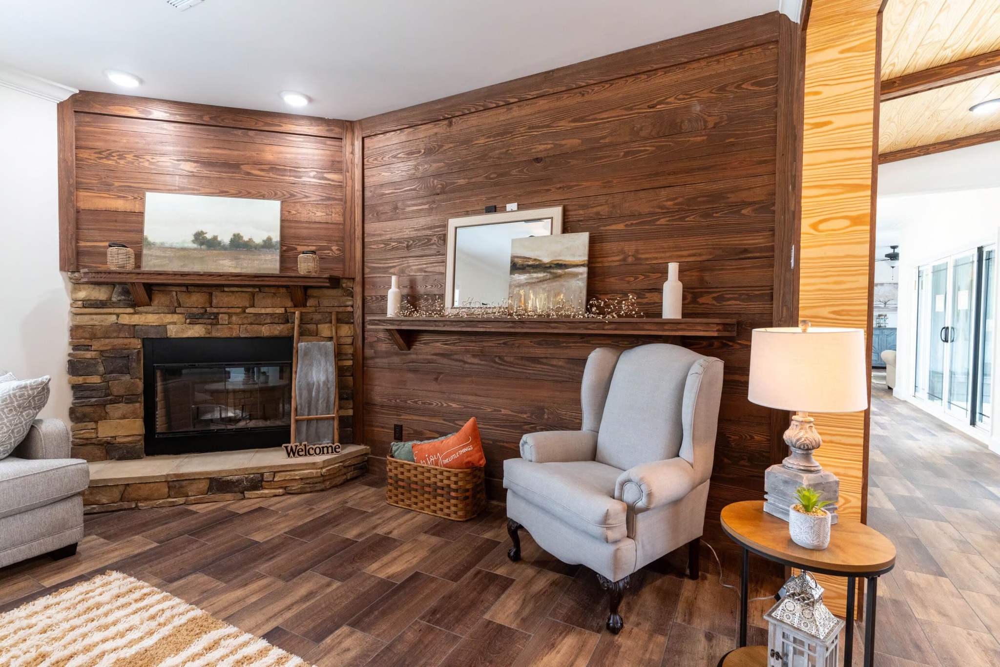 Cozy living room with wood-paneled walls, stone fireplace, and decorative mantle. Features a plush armchair, side table with lamp, and woven basket.