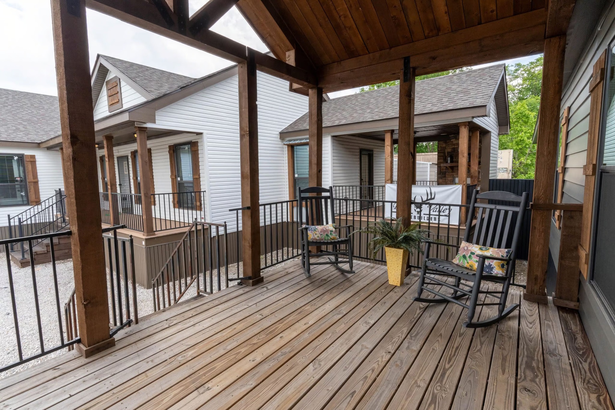 Wooden porch with two black rocking chairs and a yellow potted plant. The space is open and inviting, with views of nearby white wooden houses.
