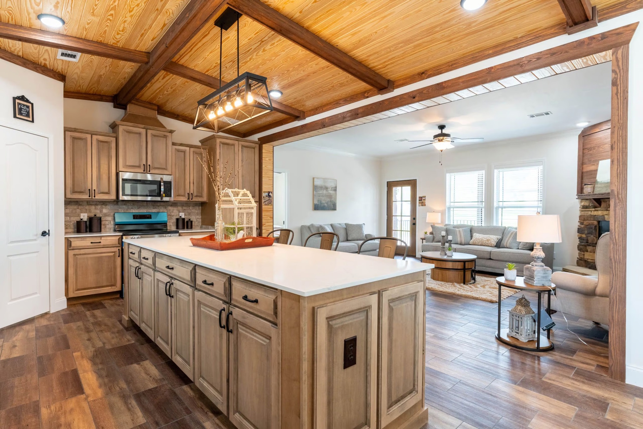 Spacious kitchen and living area with wooden ceiling beams. Kitchen features a large island, modern appliances, and rustic cabinets. Cozy living space with sofas and a ceiling fan. Bright and inviting atmosphere.