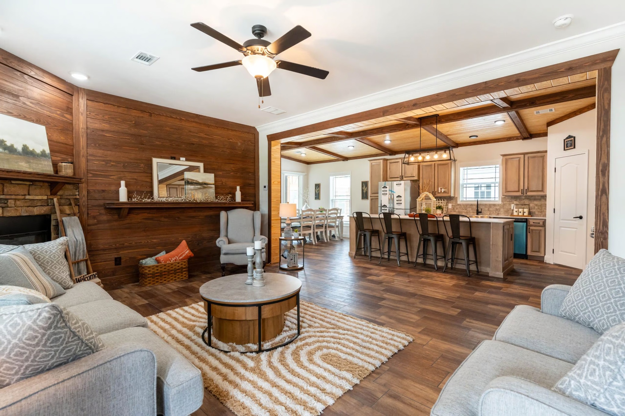 Spacious living room with wood accents, cozy gray sofas, a circular table, and rug. Open kitchen features wooden cabinets, island with stools. Warm, inviting atmosphere.