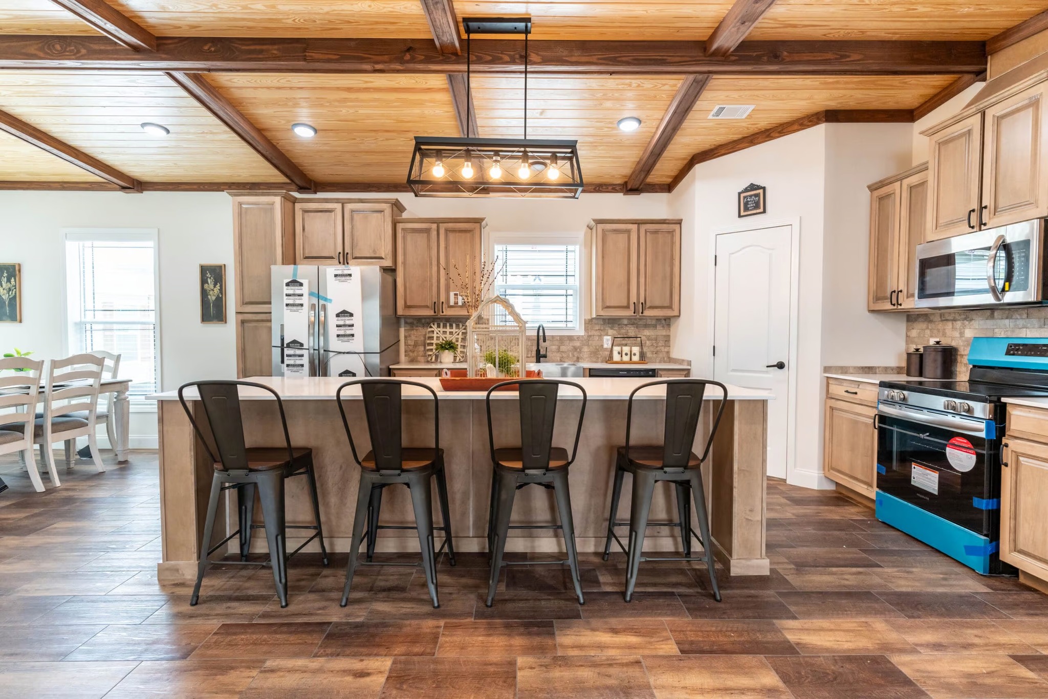 Modern kitchen with wooden cabinets, large island with four metal stools, rustic ceiling beams, and a lit pendant light. Cozy, inviting atmosphere.
