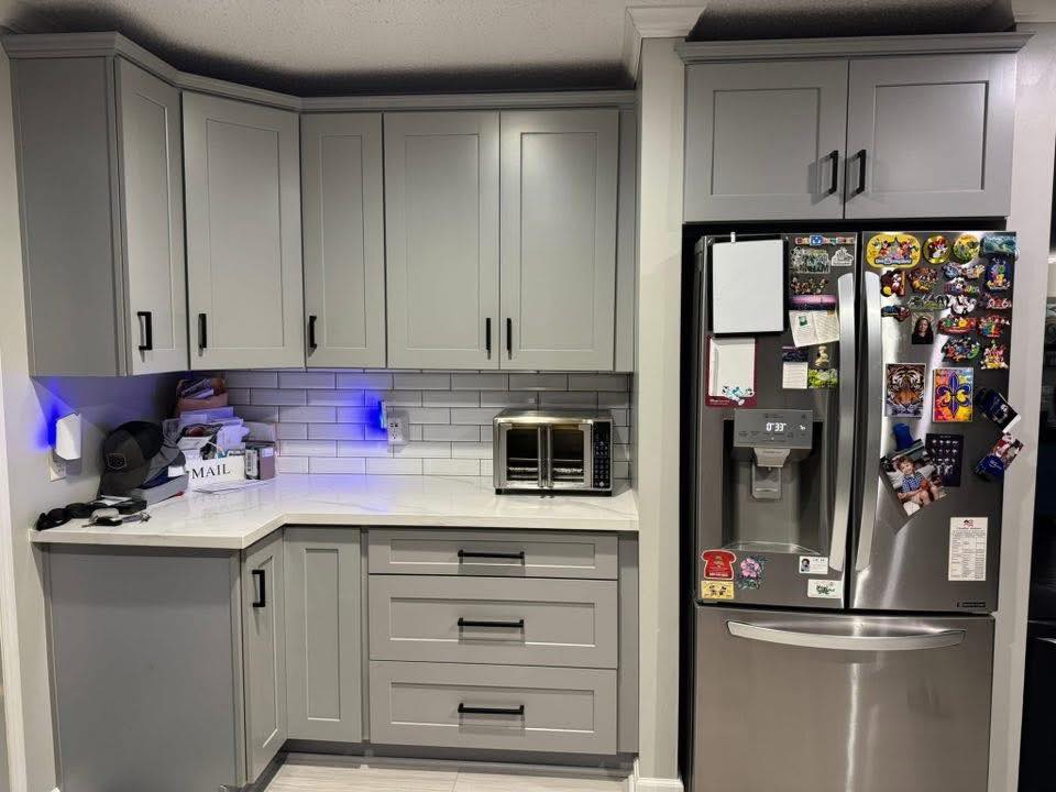 Modern kitchen with gray cabinets, white countertops, and a subway tile backsplash. A stainless steel fridge with magnets and a small toaster oven add functional decor to the cozy space.