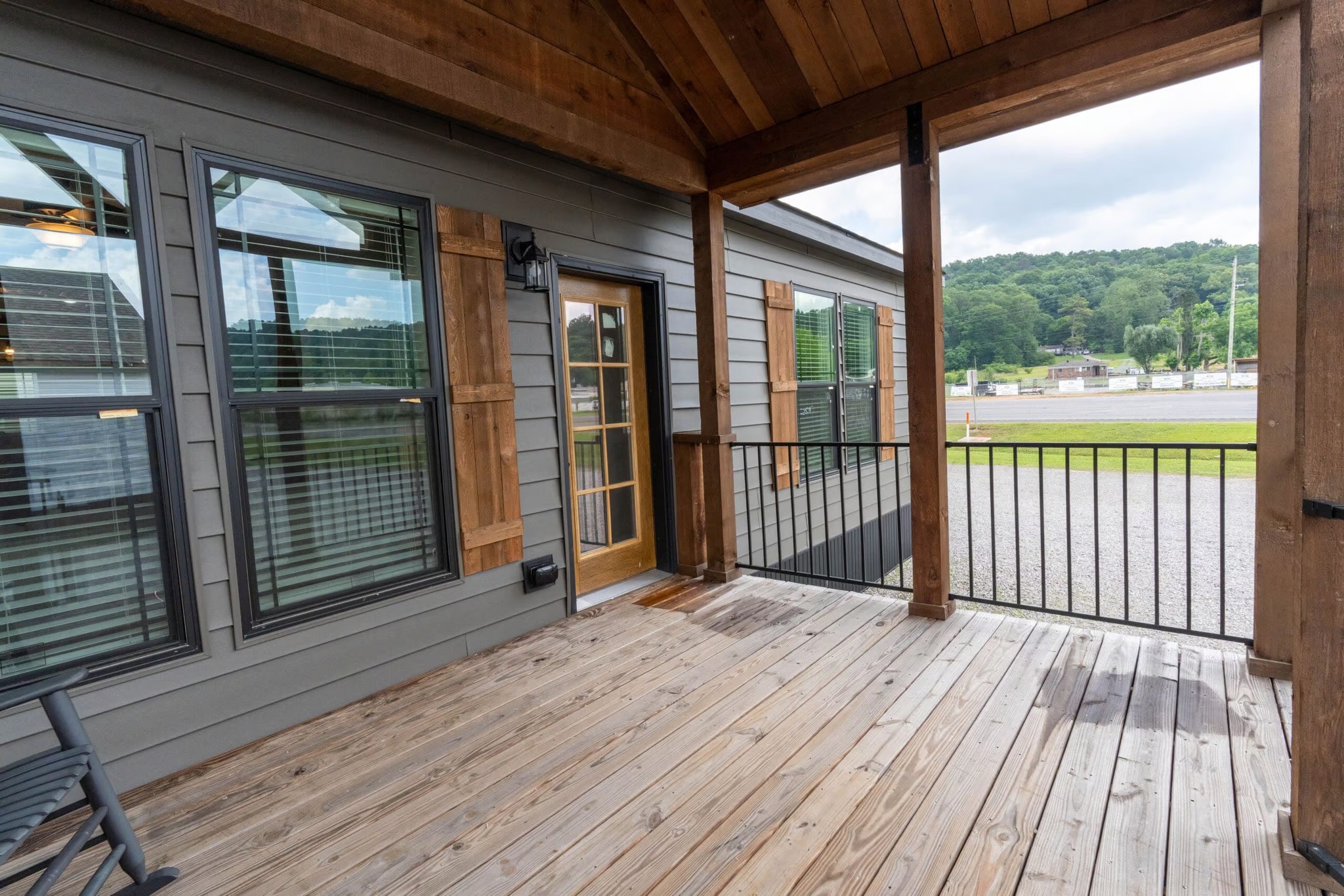 Wooden porch with a rustic feel, featuring a glass door and large windows. Overlooks a green lawn and forest area, conveying a serene atmosphere.