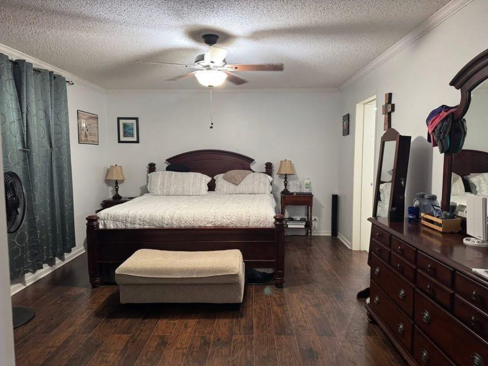 Spacious bedroom with a dark wooden bed, white bedding, and two bedside tables with lamps. A ceiling fan is above, and a dresser with a mirror is to the right.