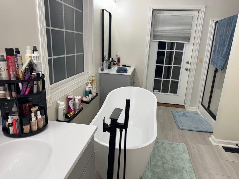Bright bathroom with a modern white bathtub, surrounded by neatly arranged toiletries. Gray floor mats and a towel hang by a door, giving a tidy and serene feel.