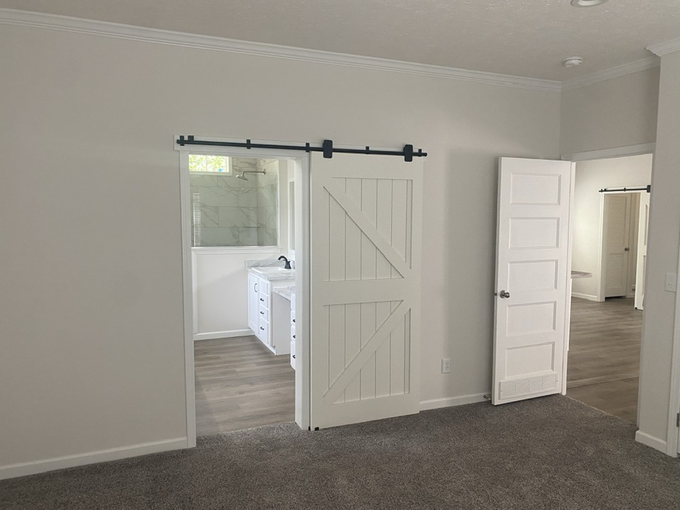 A bright, modern room featuring a barn-style sliding door leading to a bathroom. The space has light walls, carpeted and wooden floors, and a calm ambiance.