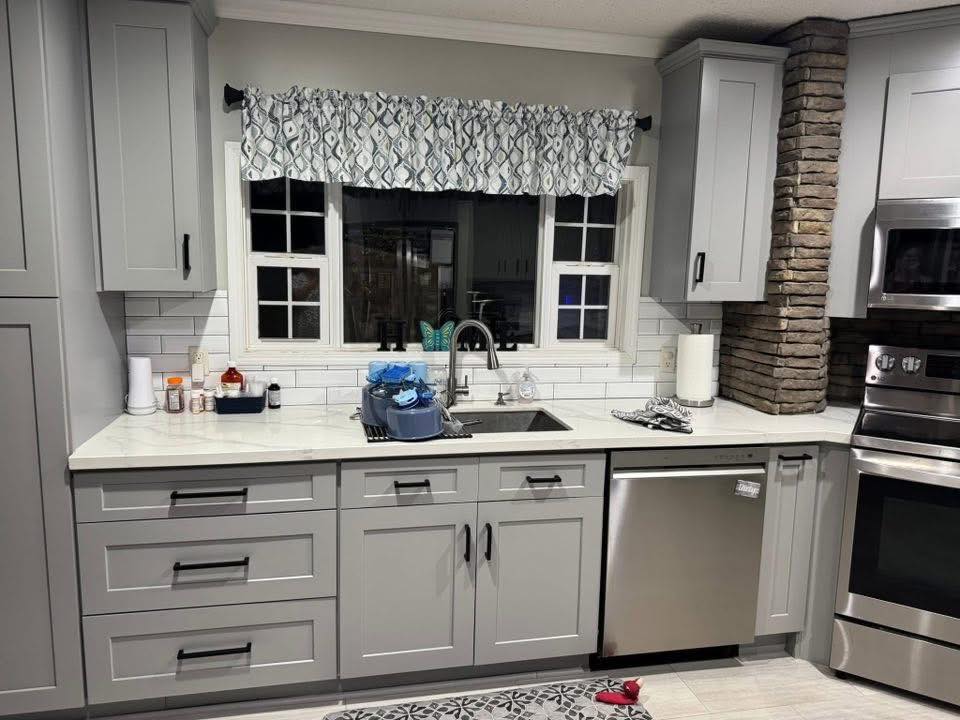 Modern kitchen with gray cabinets and stainless steel appliances. A stone accent wall adds texture. A window with patterned valance is above the sink.