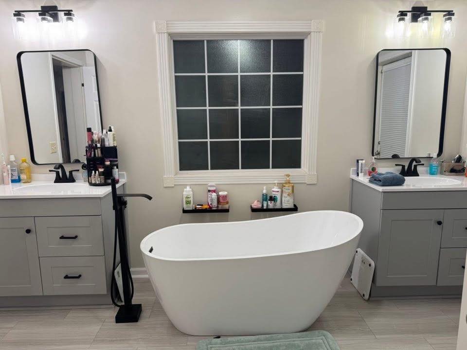 Modern bathroom with two grey vanities, rectangular mirrors, and black faucets flanking a central freestanding bathtub. A large frosted window is above the tub, and various toiletries are on the counters and window shelf. The ambiance is clean and organized.
