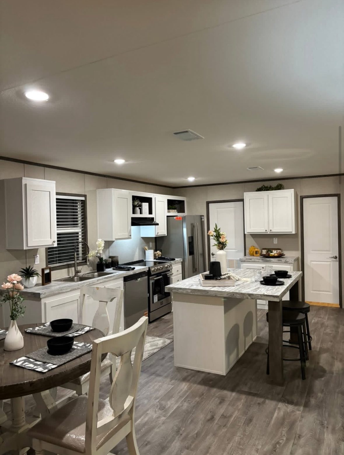 Modern kitchen with white cabinets and marble countertops featuring a central island. A round dining table is set with black dishes, under soft lighting. Cozy and inviting.