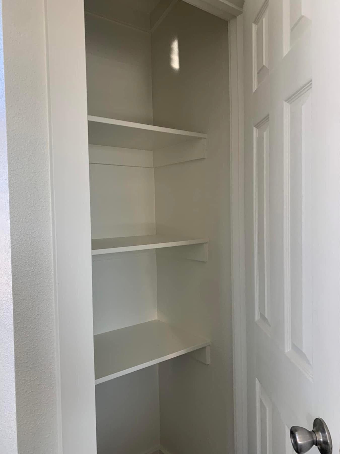 A white, open pantry closet with three empty shelves and a partially visible door on the right. The space is clean and well-lit, conveying simplicity.