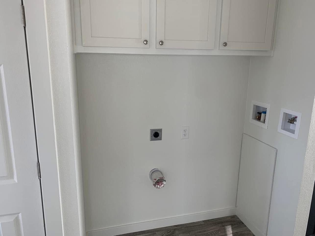 Empty laundry area with white cabinets above, utility hookups in the wall, and a gray floor. The space is freshly painted and neatly organized.