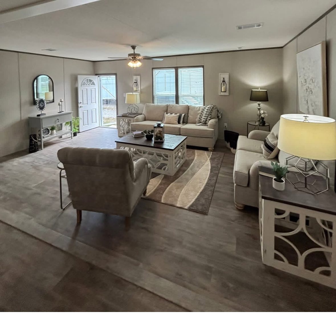 Cozy living room with beige sofas, armchair, and a coffee table on a patterned rug. Soft lighting from table lamps and a ceiling fan conveys warmth.