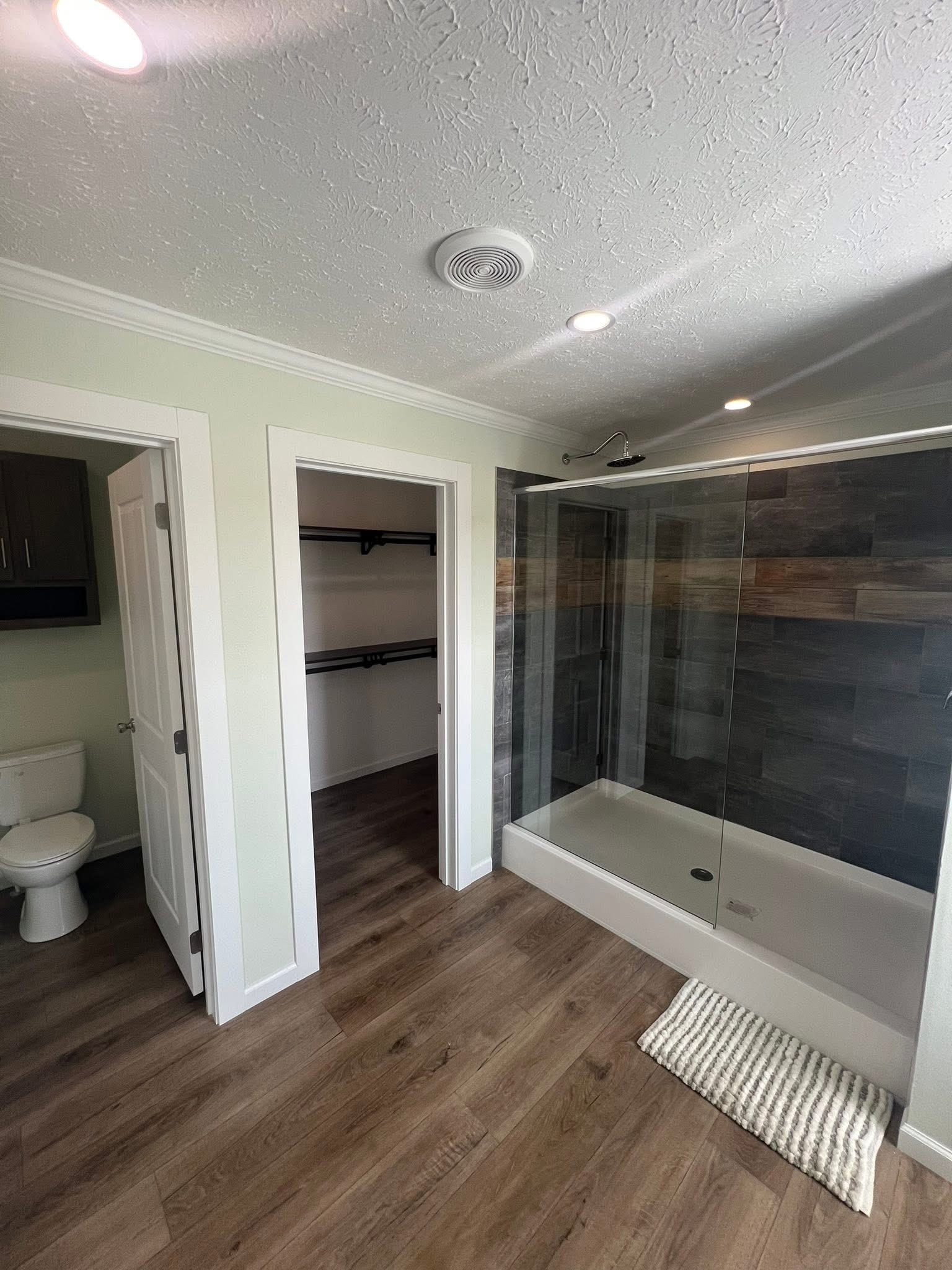 Modern bathroom with wood floors, large glass shower, textured tile wall, toilet, and open closet. Bright lighting creates a clean, spacious feel.