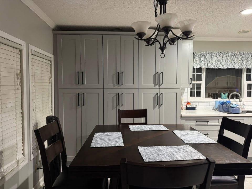 A dining area with a dark wooden table set for four, featuring light placemats. Behind, gray cabinets line the wall, and a window with a patterned curtain is above a cluttered kitchen counter. A black chandelier hangs overhead, giving the room a cozy, modern feel.