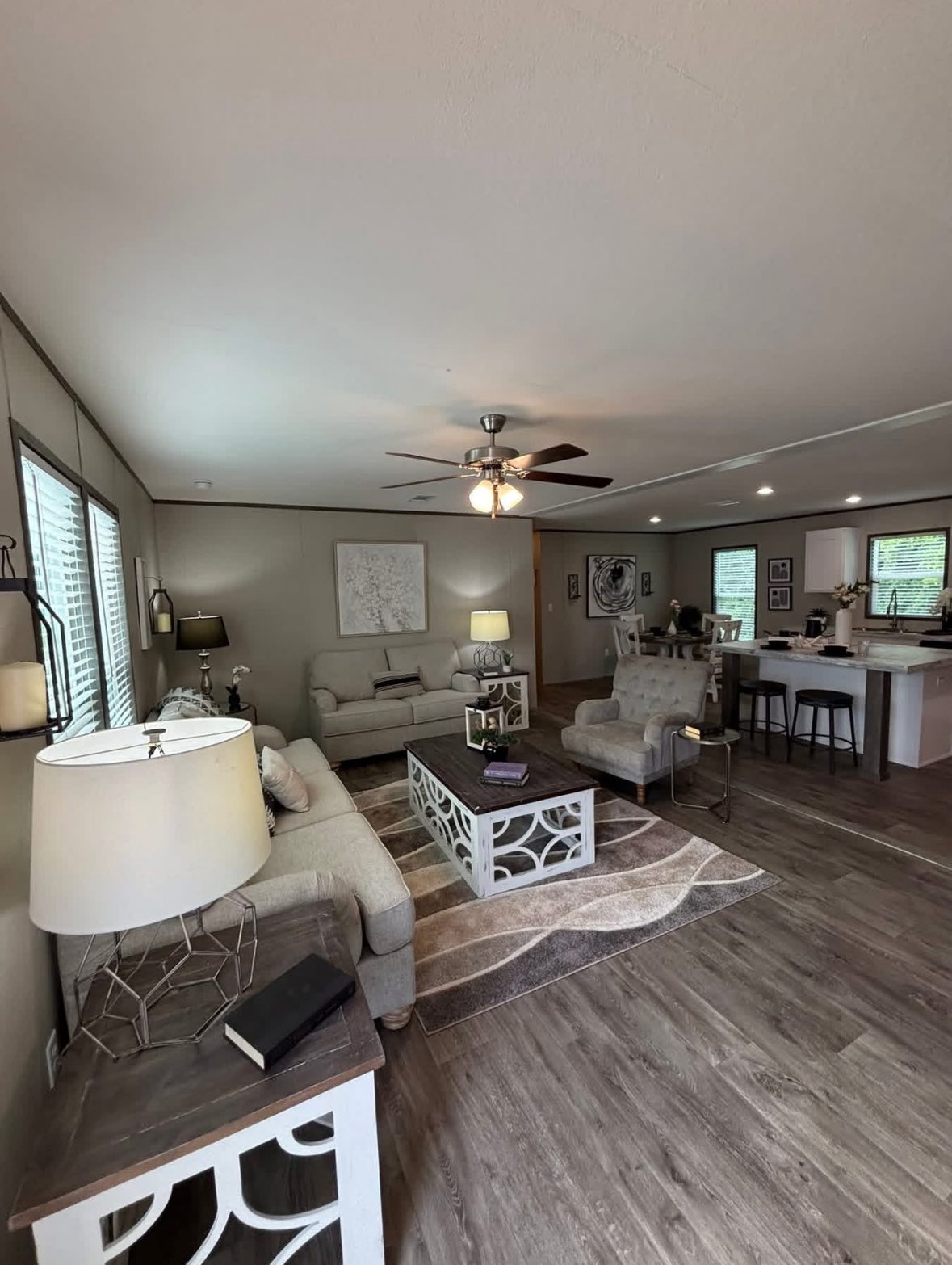 Cozy living room with neutral tones features two beige sofas, a patterned area rug, and modern furniture. A kitchen with bar stools is visible in the background.