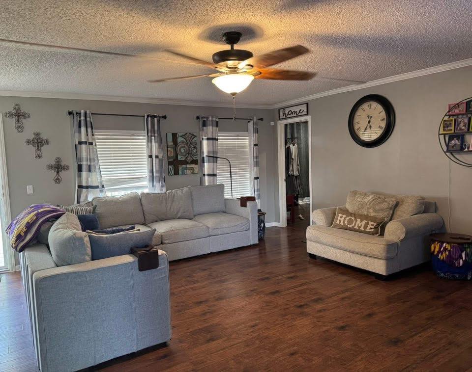 Spacious living room with wooden floor, light gray sectional sofa, and armchair. Ceiling fan, clock, and decorative crosses on the wall. Cozy ambiance.