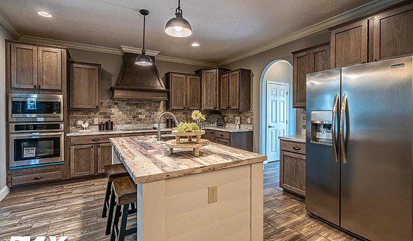 A rustic kitchen with wooden cabinets, stone backsplash, and stainless steel appliances. A large island with a mix of wood tones stands center, under pendant lighting.