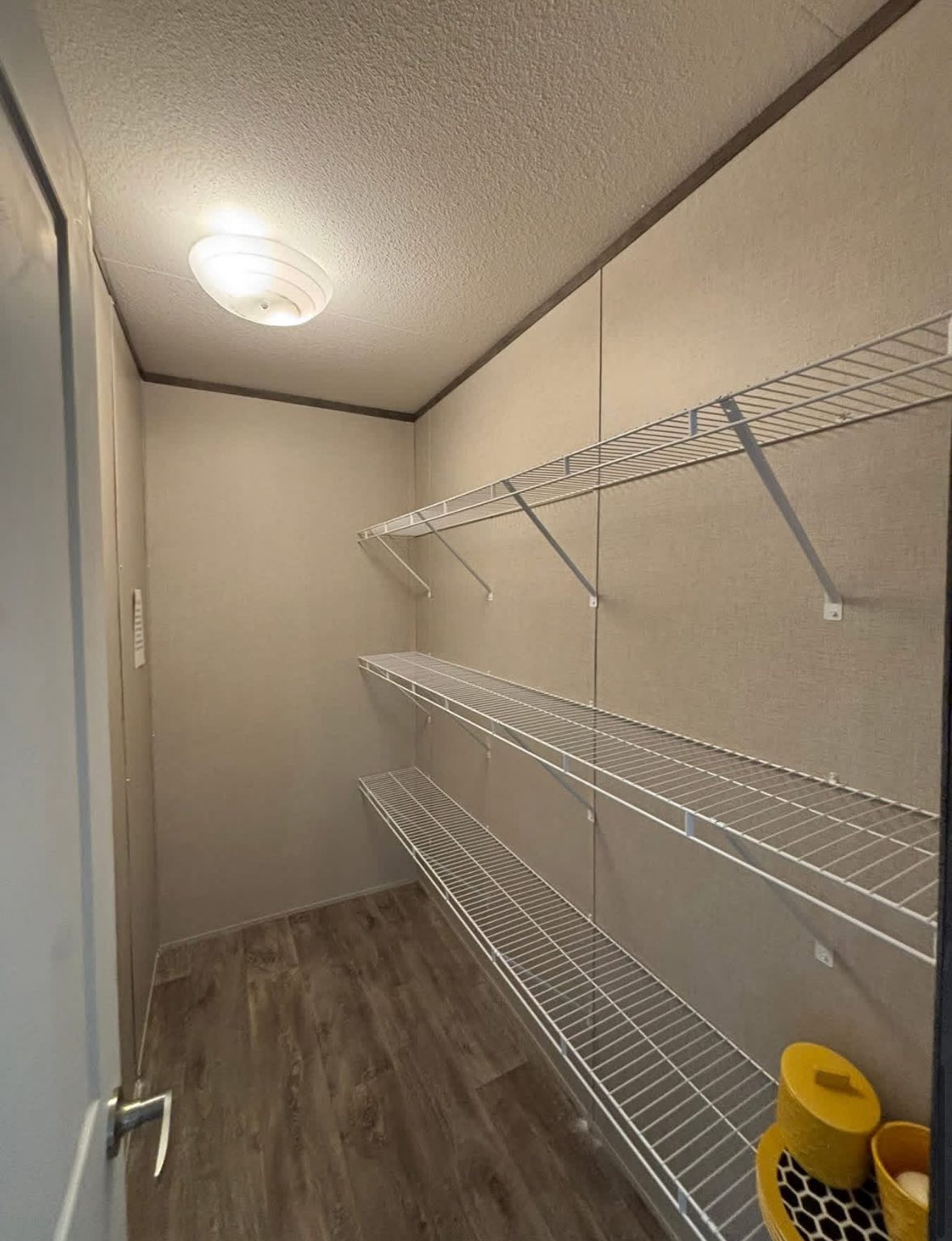 Narrow, empty closet with three metal wire shelves on the right wall. Overhead light casts warm glow on the wooden floor, creating a clean, minimalist feel.