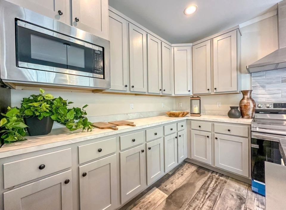 Modern kitchen with light gray cabinetry, stainless steel microwave, and stove. Decor includes plants and vases on a marble countertop. Warm, inviting tone.