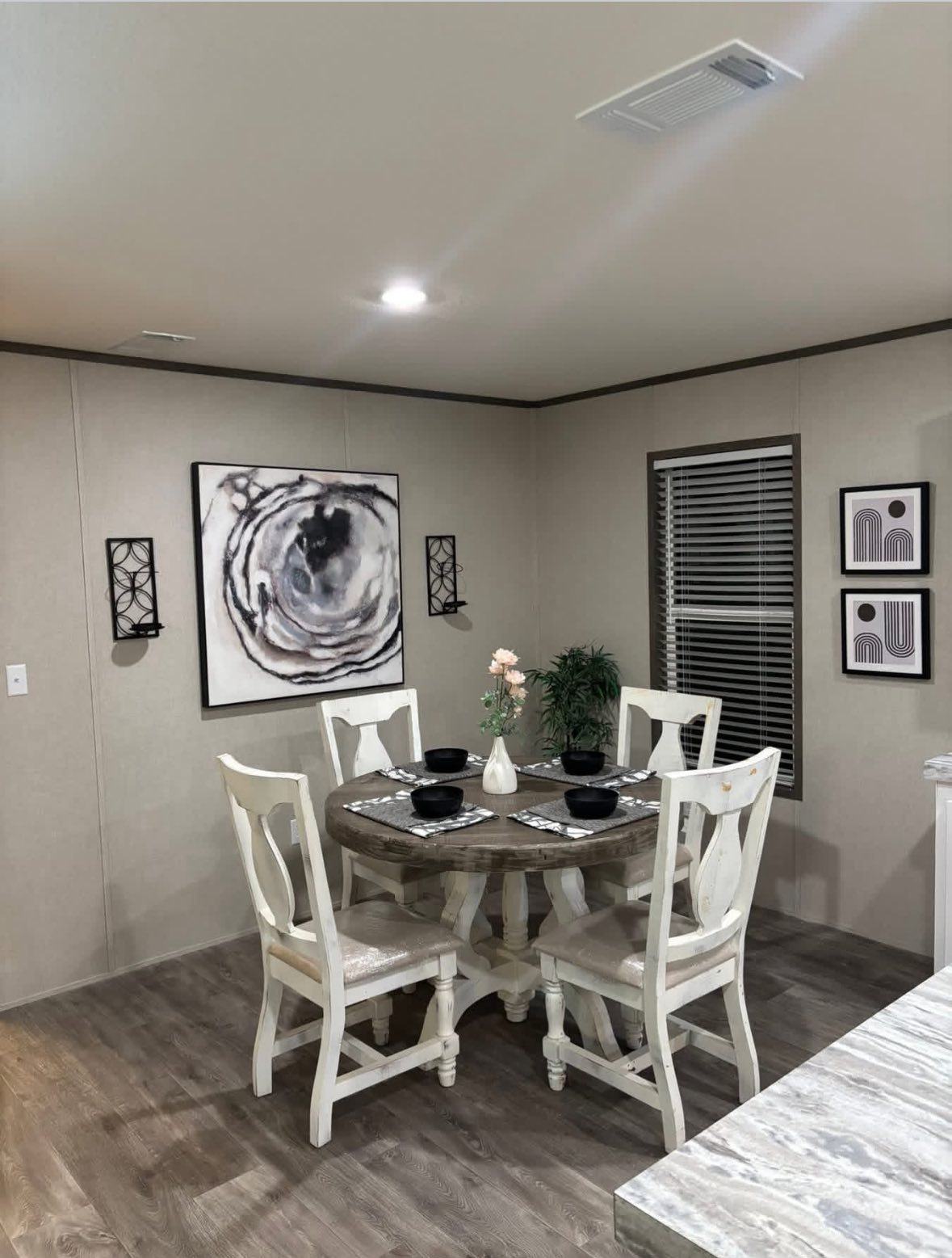 Dining room with a rustic wooden table set for four, featuring black dishes. Abstract art and two black sconce lights adorn the beige walls.